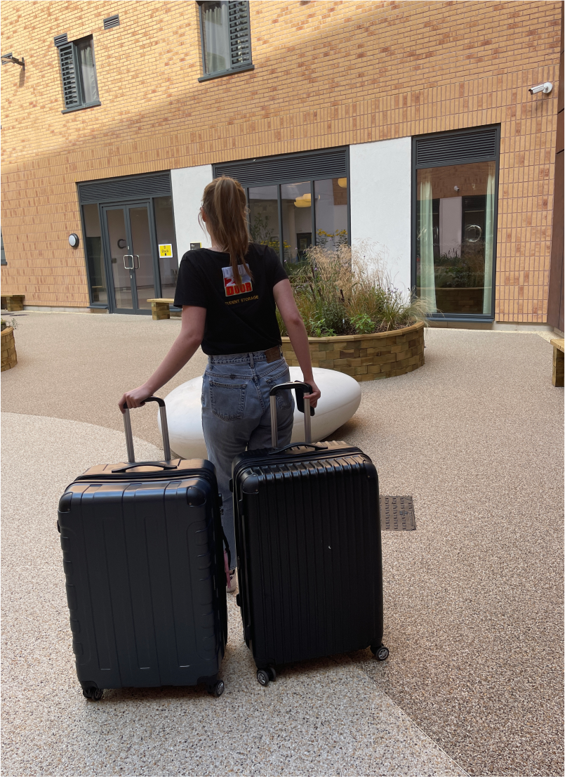 The image shows a female Door2door employee, wearing a Door2door logo black top, delivering 2 suitcases to a student accommodation