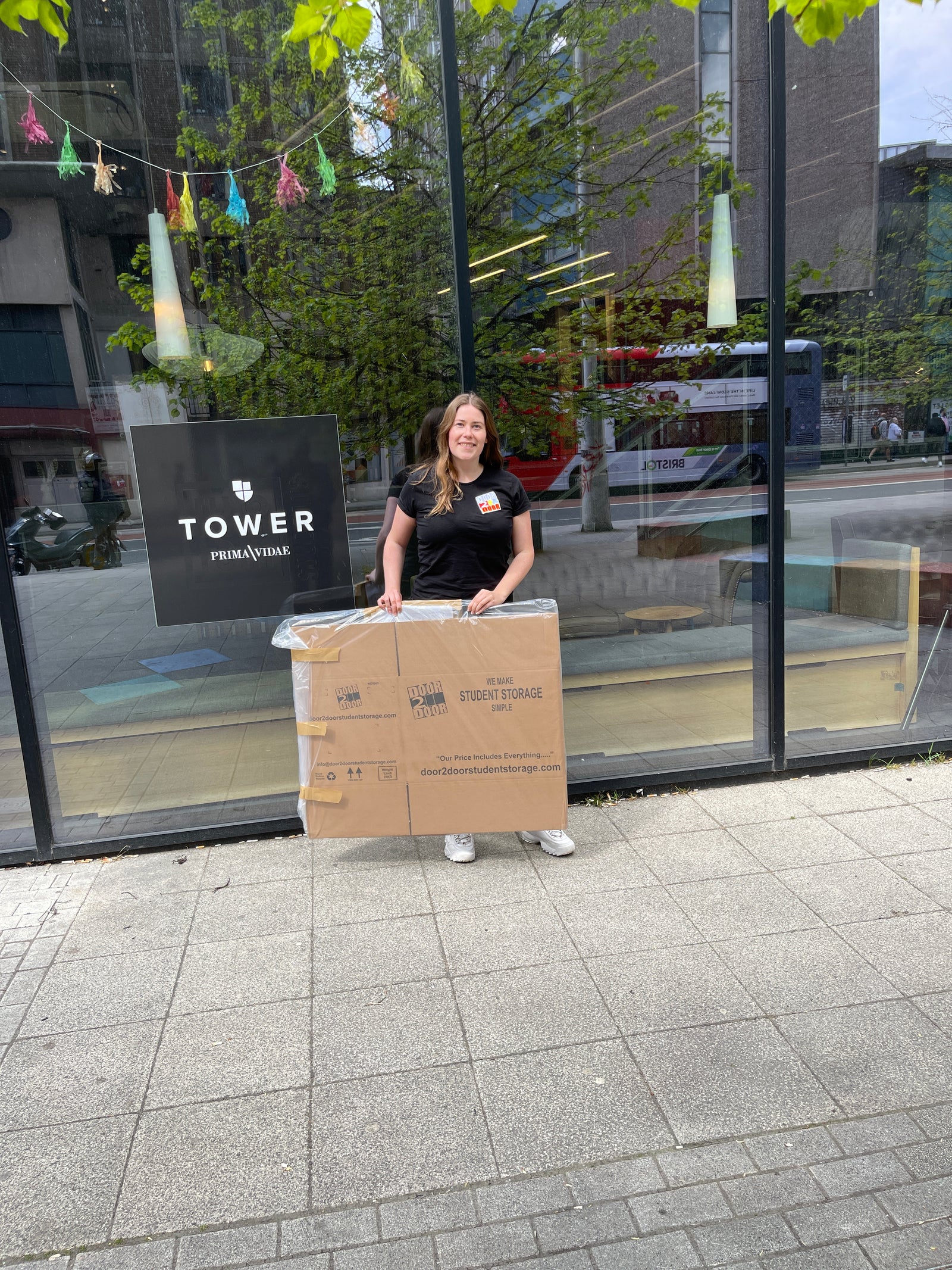 The image shows a female Door2door employee wearing a door2door logo black top, delivering empty Door2door boxes to a student accommodation