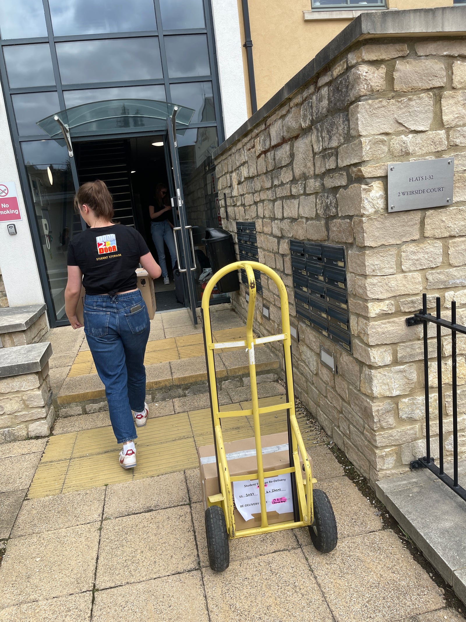 The image shows a female Door2door employee carrying a cardboard box into a student accommodation recpetion room. the image also shows a yeloow sacktruck with another cardboard box on.