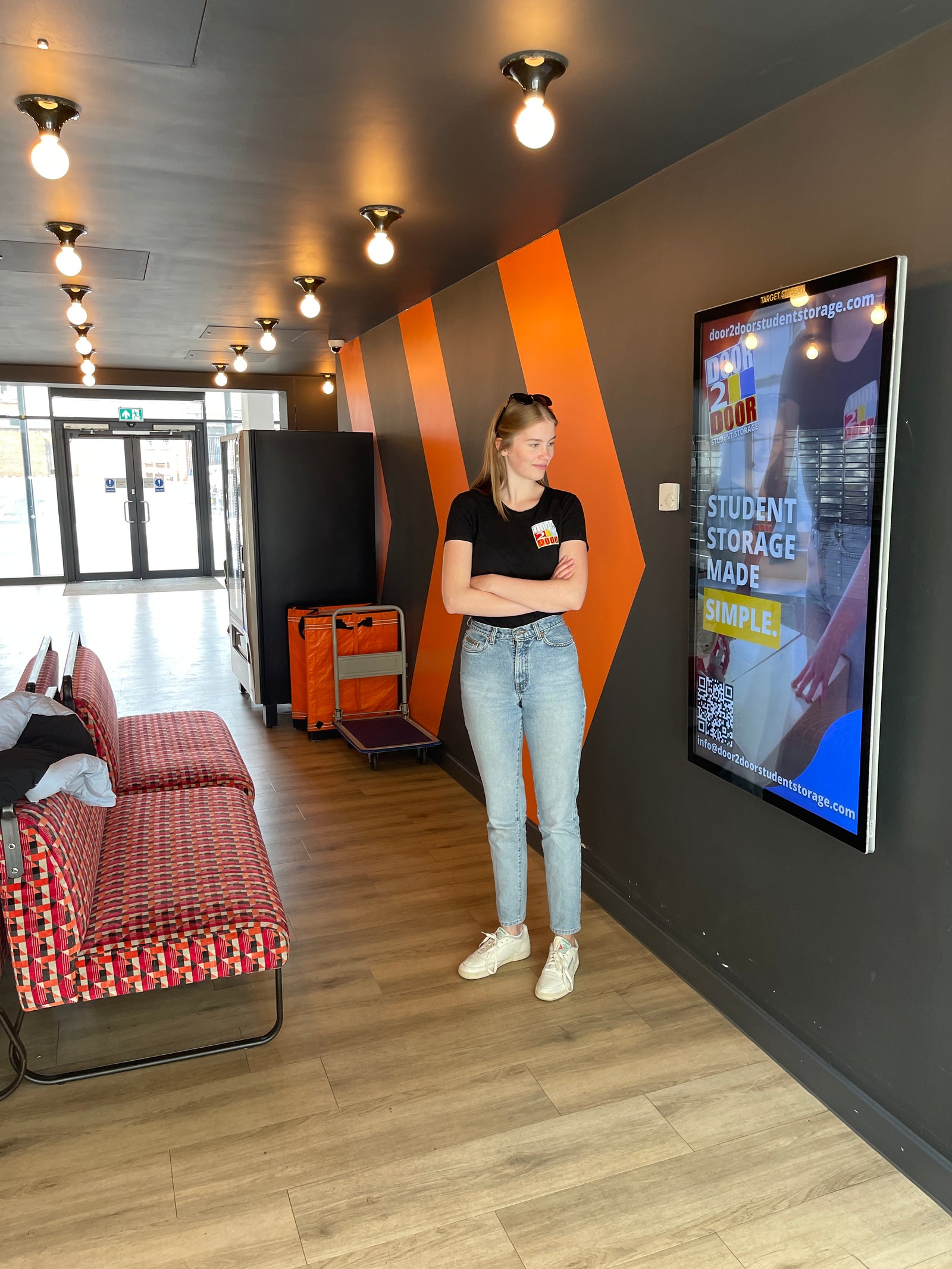The image shows a female Door2door employee wearing a Door2door logo black top, standing in front of a screen showing a Door2door ad, inside a student accommodation