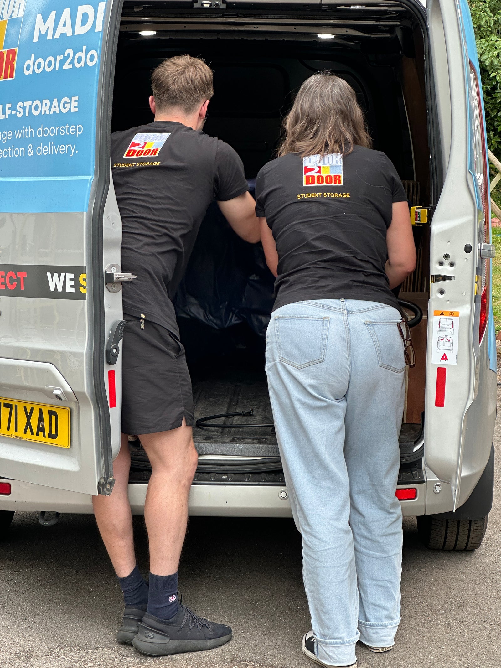 The image shows two Door2door employees, one femlae and one male, both wearing Door2door logo black tops. The Door2door employees leaning in the rear end of a Door2door van 